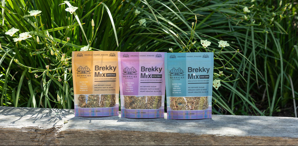Three packages of Brekky Mix on a wooden surface with greenery in the background