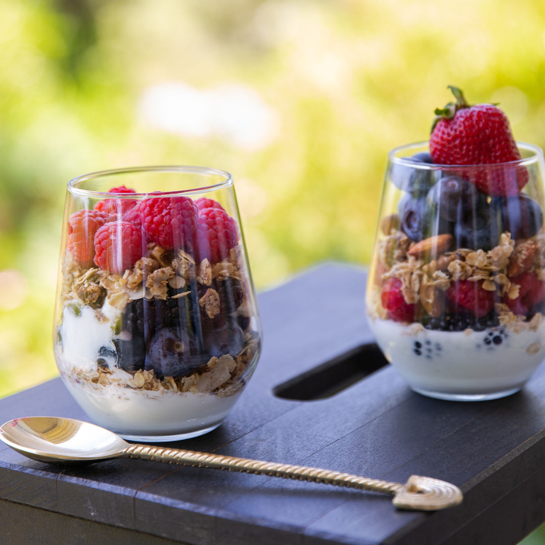 Two glasses of yogurt with low sugar granola and berries on a wooden surface outdoors.